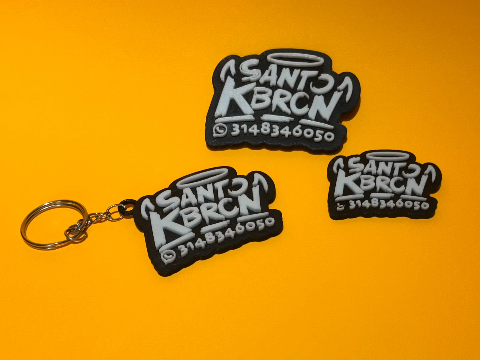 Tres piezas con el logo de un negocio en relieve blanco sobre negro sobre fondo naranja, una de ellas con la argolla metálica de llavero puesta — Llaveros e imanes de Solid Lab 3D, Colombia