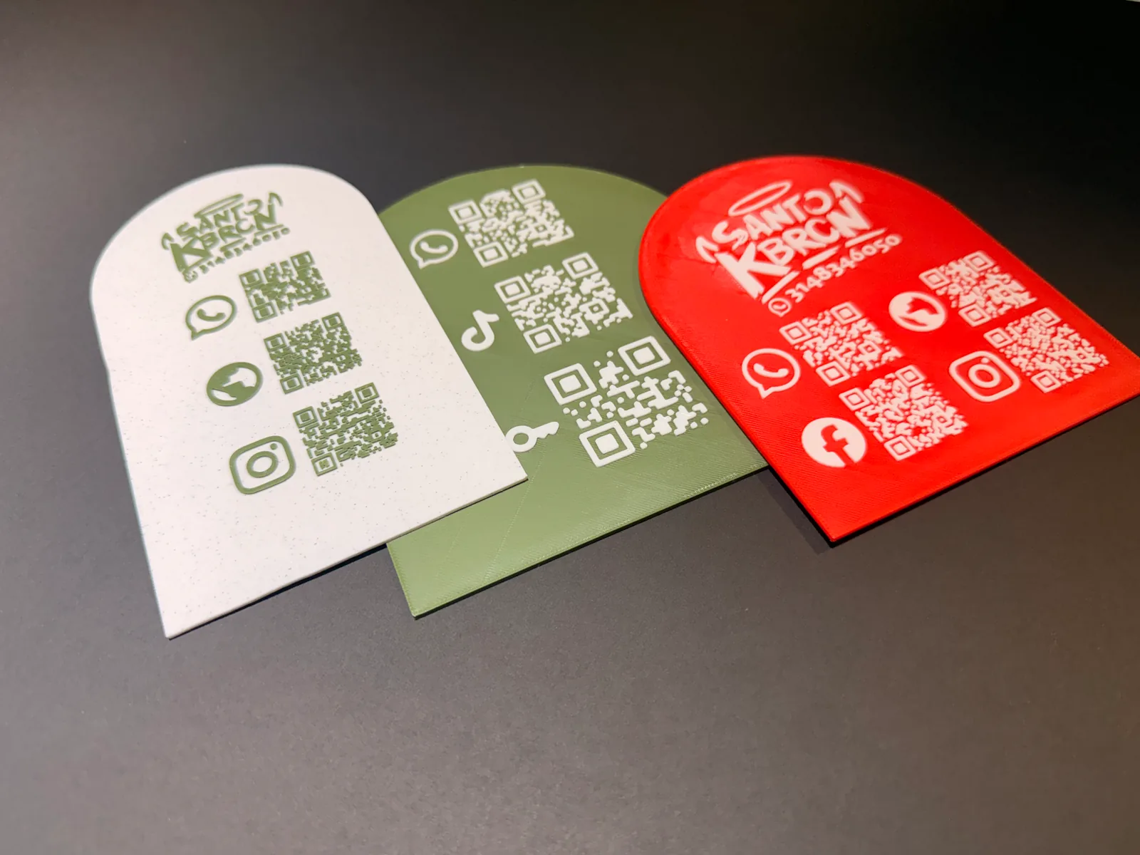 Tres Placas Portátiles QR en crema, verde oliva y roja inclinadas sobre fondo negro, con los códigos QR de WhatsApp, sitio web y redes sociales en relieve — Placa Portátil QR de Solid Lab 3D, Colombia